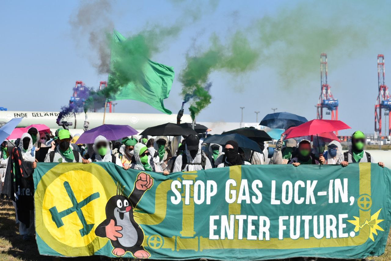 Transpi 'stop gas lock-in, enter future' mit Bunten Fahnen und Rauch im Hintergrund